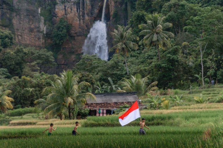 Tiga anak laki-laki bermain dengan bendera Indonesia dan air terjun di latar belakang