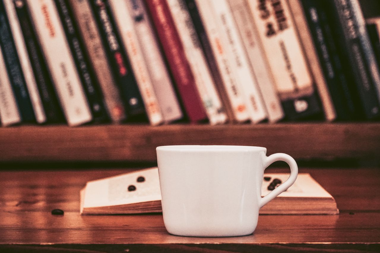 cangkir-kopi-putih-dan-buku-terbuka-di-meja-kayu-dan-rak-perpustakaan